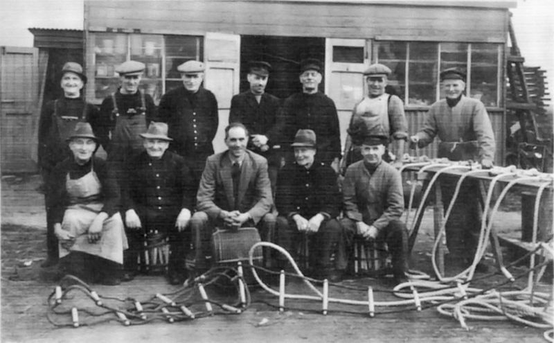  Ernest Appleton (front row on the left) amongst a group of workers employed by Gowens in Tollesbury during WW2. They made rope ladders, scramble nets etc.

Back row L to R. 1. Jim Appleton, 2. Jimmy Lewis, 3. Will Frost, 4. Dick Holder, 5. Charlie Pettican, 6. Dick Lewis, 7. Silas South.

Front row L to R. 1. Ernie Appleton (twin of Jim), 2. Bob Appleton (brother of Jim and Ernie), 3. Ken ...
Cat1 Tollesbury-->People Cat2 People-->Fishermen and Seamen Cat3 Tollesbury-->Shops and Businesses
