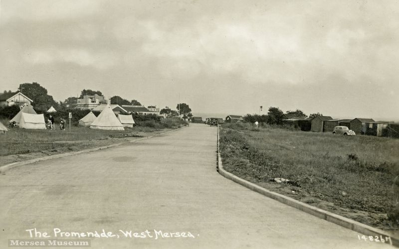  The Esplanade near the bottom of Empress Avenue. Camping on the grass on the left. Fairhaven Avenue beyond with the house attached to Fairhaven Cafeteria just being built. It did not survive many years. The toilet block this side of Fairhaven has not yet been built.

See GEO_001_002 for the back of the postcard, which says This go up past Grays [who lived in Broomhills Road] to Sea View ...
Cat1 Mersea-->Road Scenes